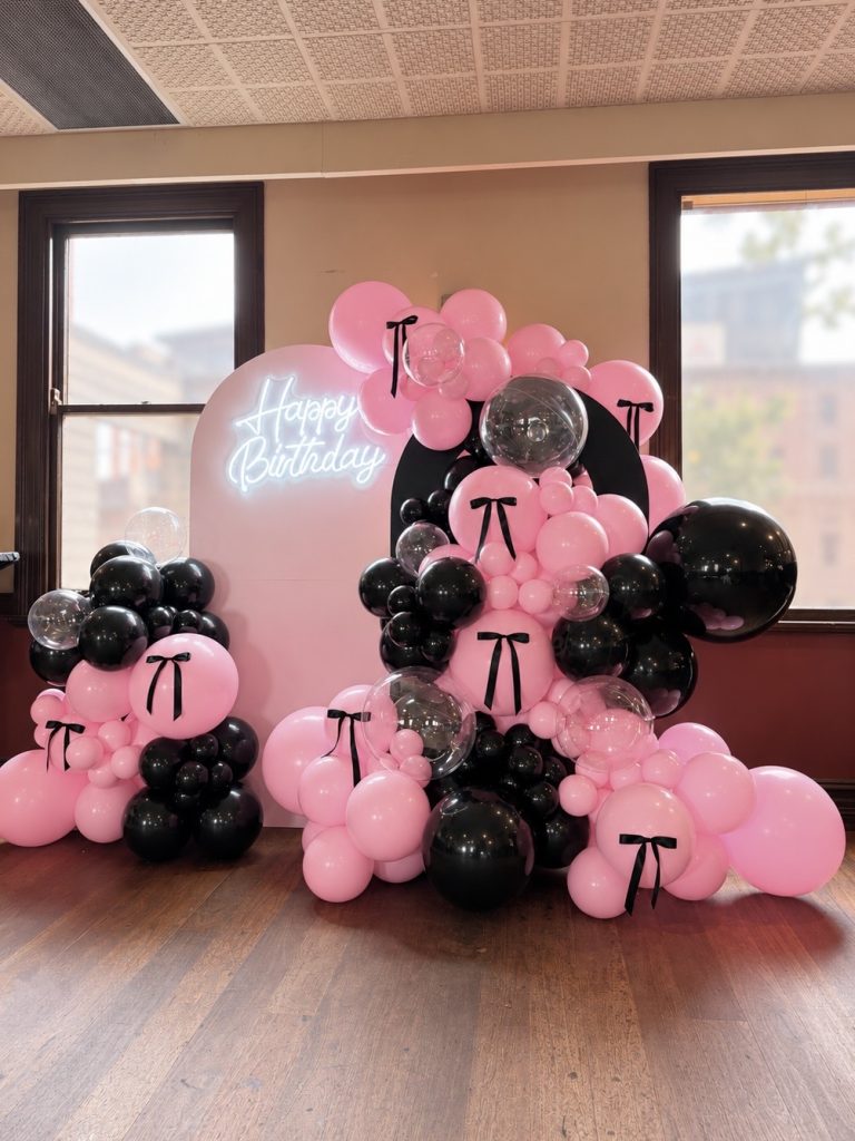 Elegant pink and black birthday balloon setup featuring clear bubble balloons, decorative black bows, layered circular backdrops and glowing Happy Birthday sign.