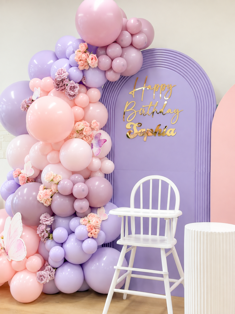 Pastel pink and lavender balloon arrangement with floral accents, a personalised purple first birthday backdrop and white baby chair.