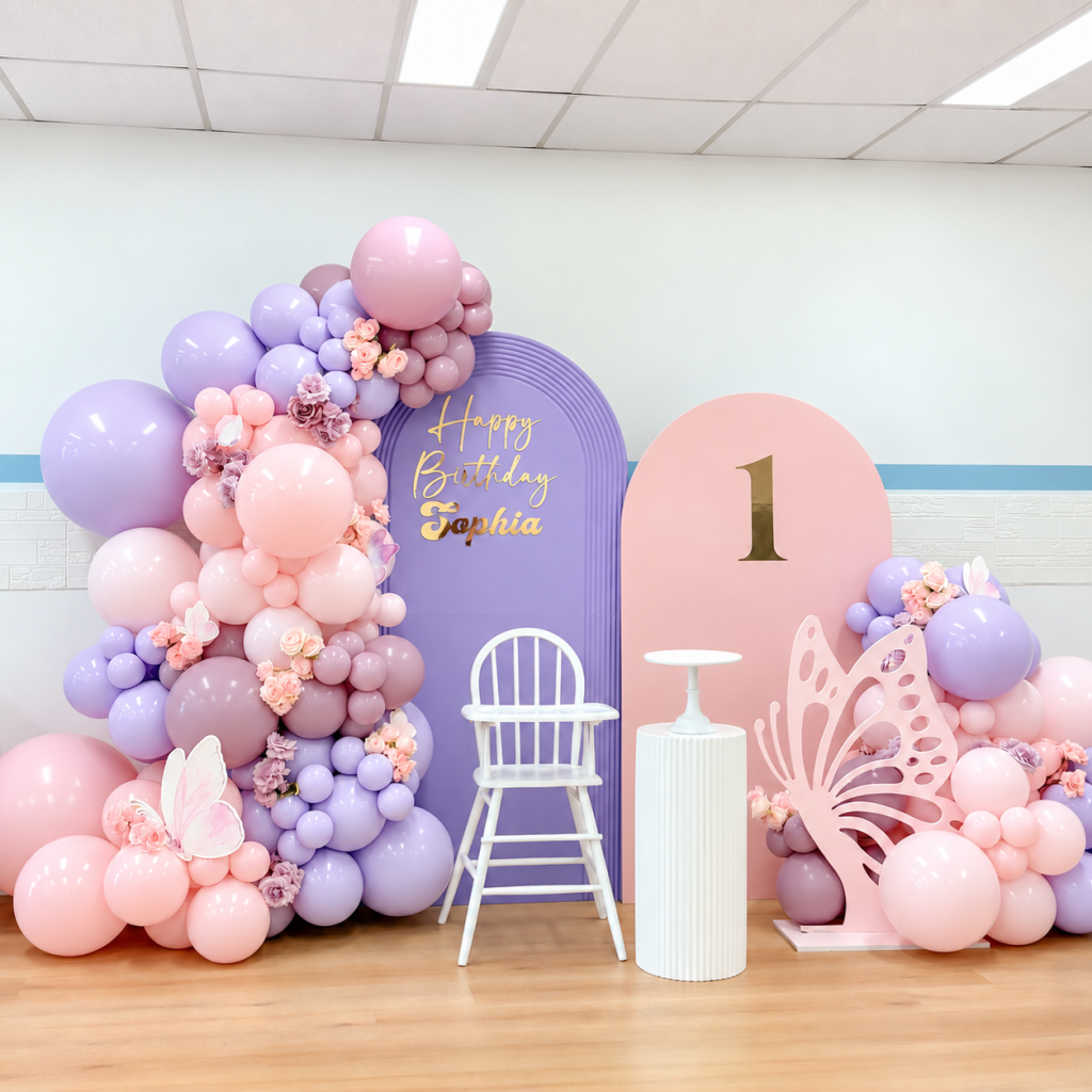 Large decorative pink butterfly beside pastel pink, purple and white balloons at a first birthday celebration.