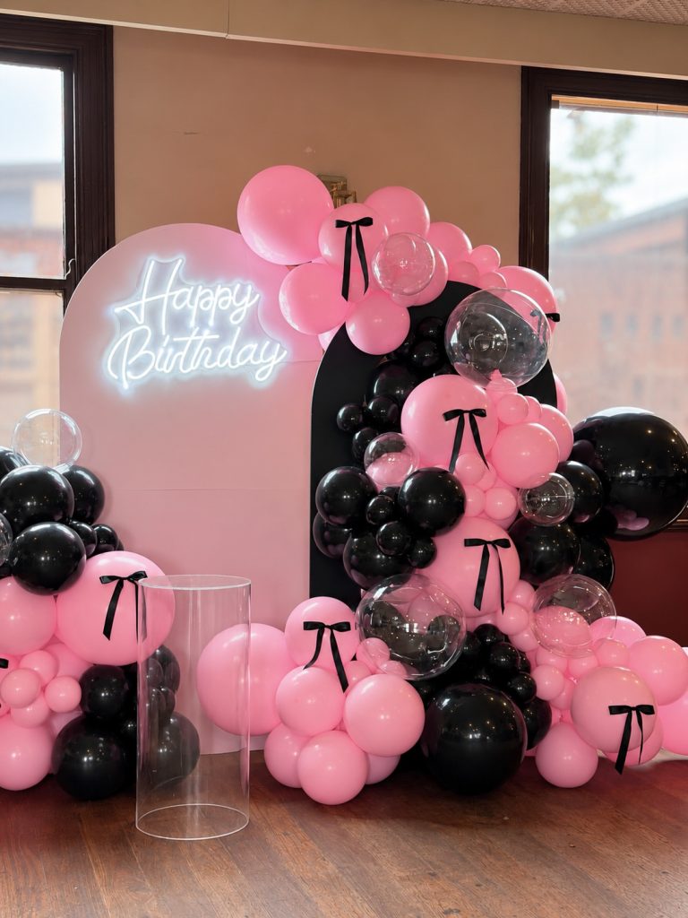 Black and Pink Birthday Balloon Decoration with illuminated Happy Birthday sign, pink backdrop, black bow accents and organic balloon garlands.